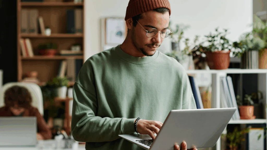 A young man in a beanie and glasses uses a laptop to learn how to make money with bumper stickers.