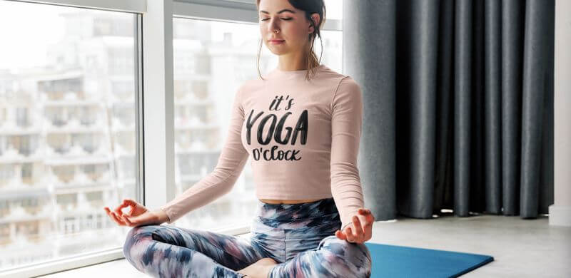 A woman is sitting on a yoga mat wearing a long-sleeve shirt that says It’s yoga o’clock.
