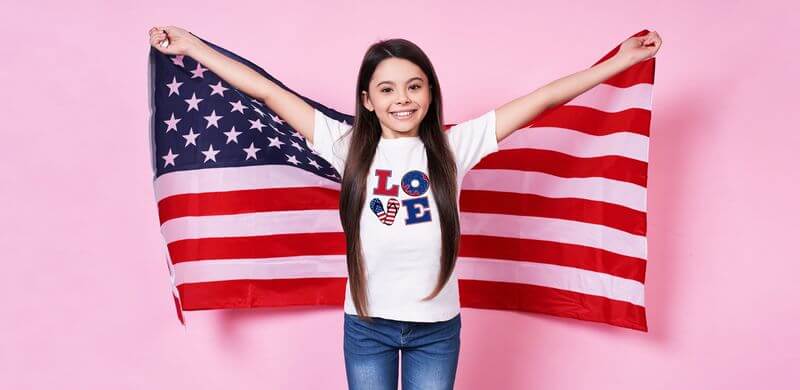 A child is wearing a t-shirt that spells LOVE while holding the United States flag behind her.