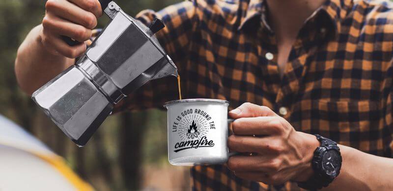 A man pours coffee into an enamel mug that says Life is good around the campfire. It features a minimalistic illustration of a campfire.