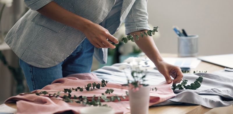 A person styling a scene for a photoshoot. They place green plant decorations on top of some t-shirts.