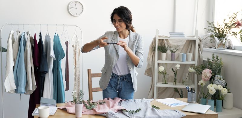 In a casual office, a woman takes a photo of her products on a table with her phone.