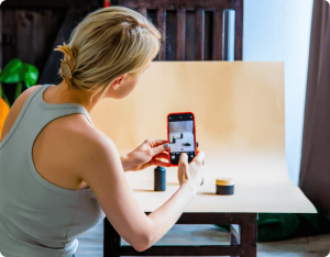 A woman takes a photo of her products on a beige paper background with her phone.