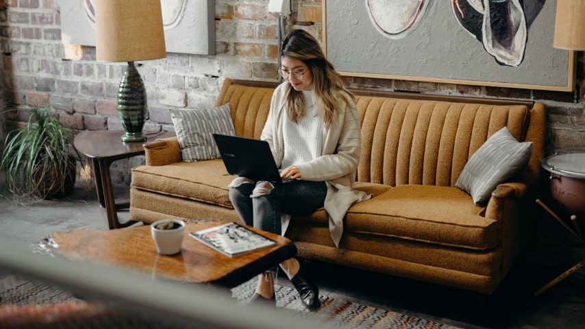 A woman works on her laptop while sitting on a couch.
