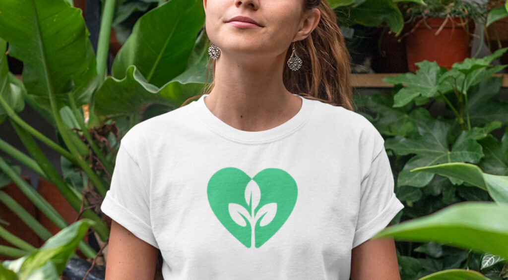 A woman in a green heart t-shirt stands in front of lush green plants, smiling and enjoying the natural surroundings.
