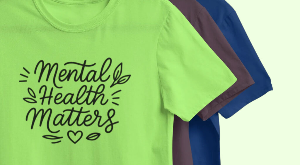 A lime-green T-shirt features the words "Mental Health Matters" in playful, black script with decorative leaves and a heart. Behind are mauve and blue shirts.