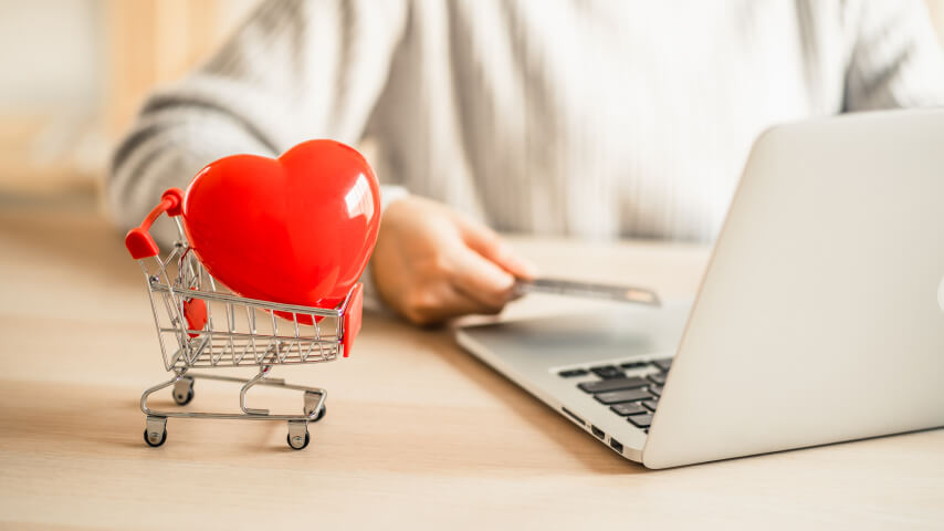 A person is sitting by the desk in front of an open laptop computer; they are holding a credit card. In front of them is a miniature shopping heart carrying a red plastic heart.