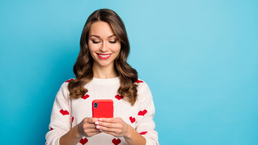 Standing against a blue backdrop is a dark-haired woman wearing a white knit sweater with red hearts. She's smiling and holding a red smartphone.