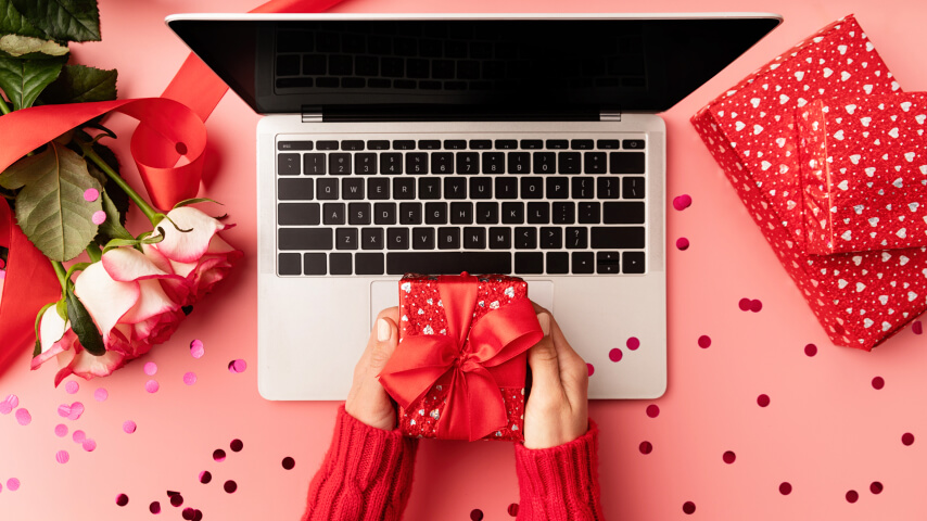 Open laptop set on a romantically decorated table and woman's hands are holding a nicely wrapped Valentine's Day gift above it.