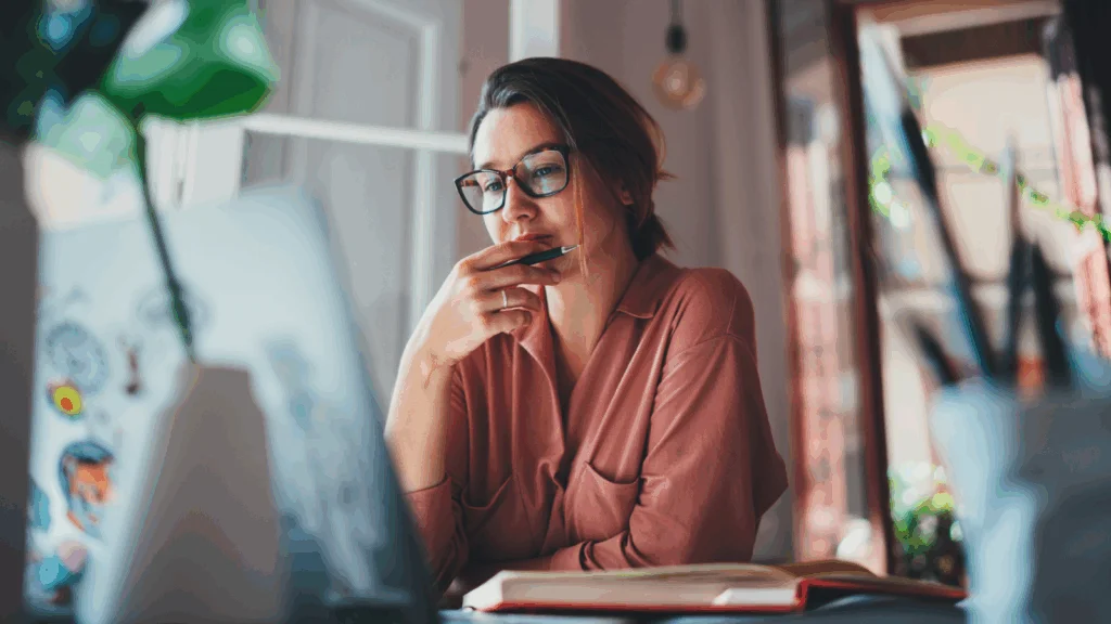 A woman wearing glasses sits at a desk, focused on her laptop while working.