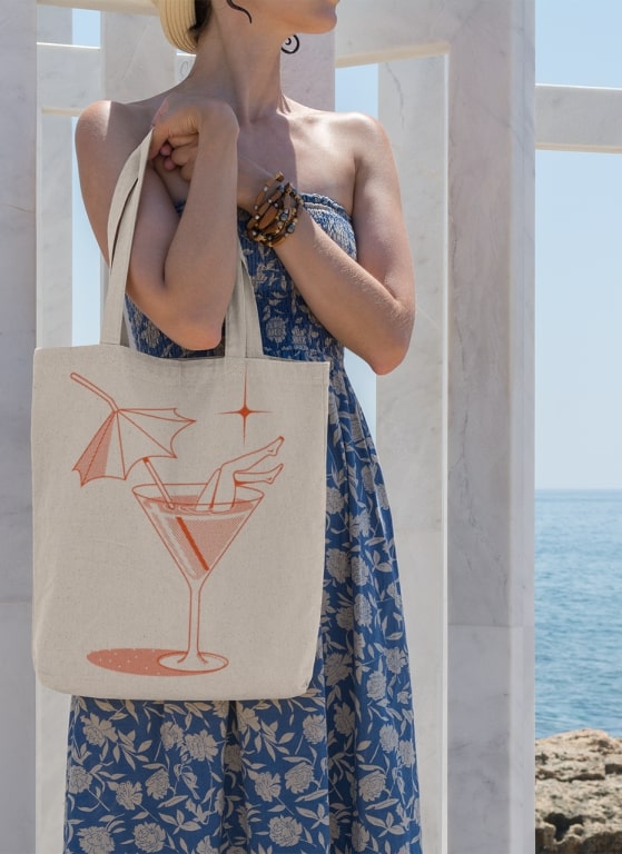 Woman near a beach, holding a tote bag with a stylized martini glass design printed on it.