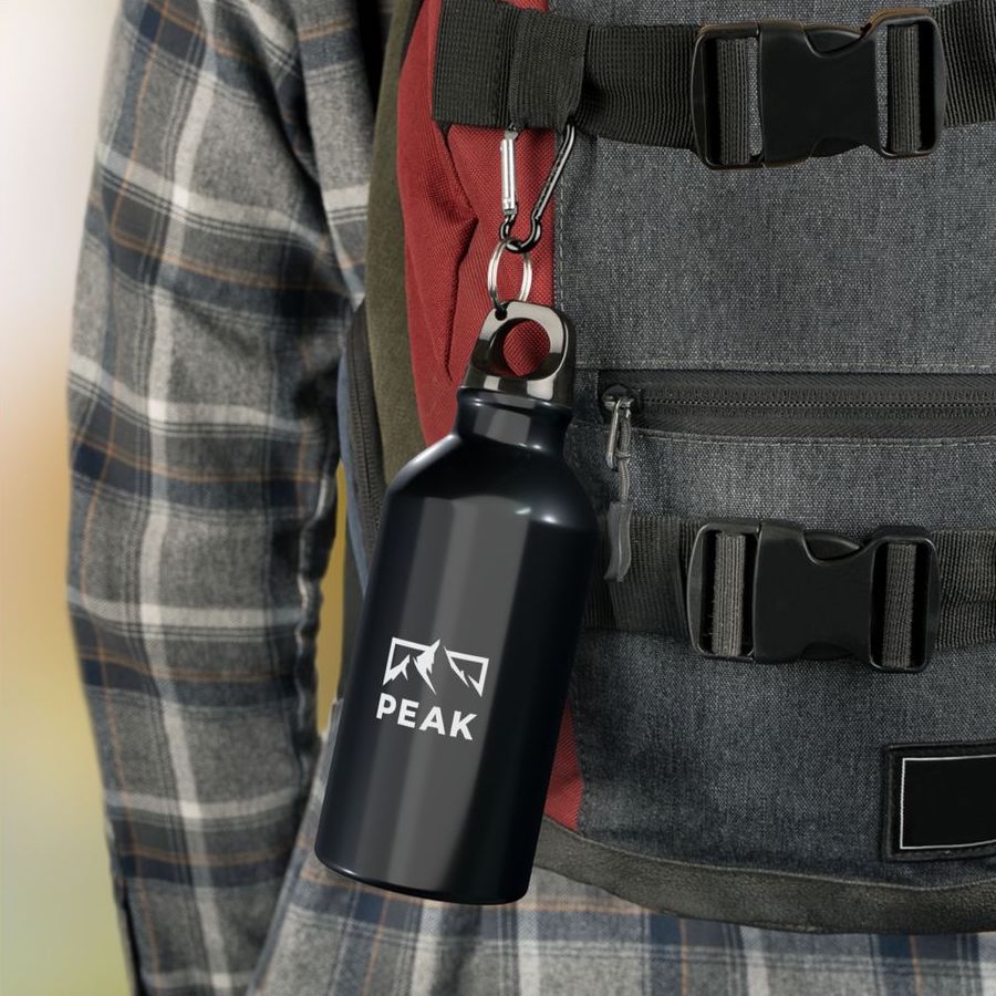 A black water bottle hooked to a hiker's backpack with a design of a mountain silhouette and the word “Peak” printed on.