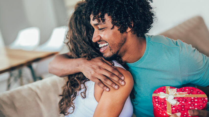 A dark-skinned couple is sitting on a couch, hugging and smiling; the gentleman is holding a red heart-shaped box of chocolates.