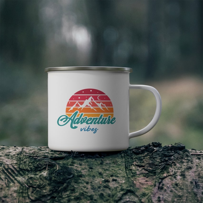 A white enamel mug standing on a log with a custom design that has a picture of mountains and words "Adventure Vibes".