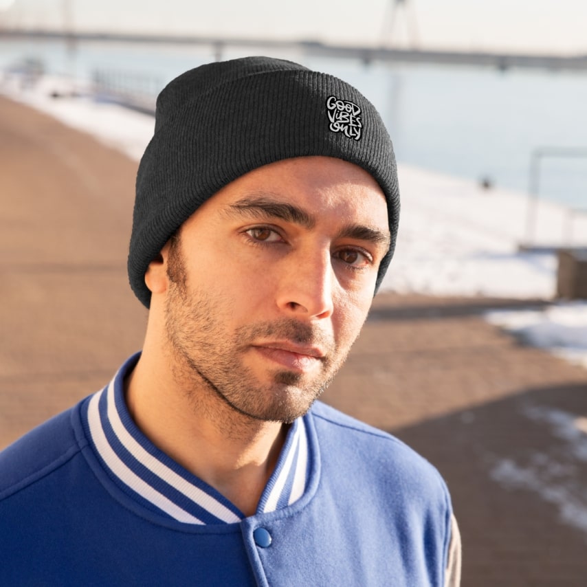 A man wearing a blue jacket and a black beanie hat with a custom design that says "Good Vibes Only".