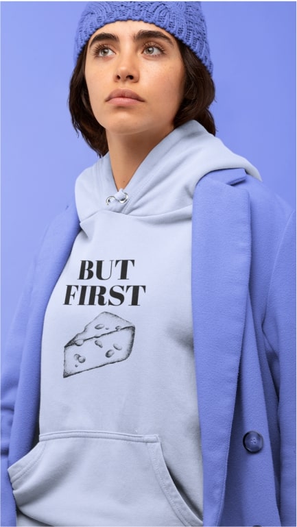 A woman posing in a blue blazer, beanie, and a grey hoodie that says "But first" and a picture of cheese