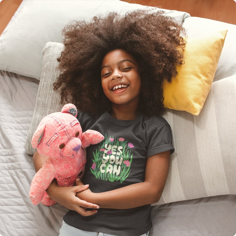 A smiling kid laying in pillows, holding a pink teddy bear, and wearing a custom shirt that says "yes we can"