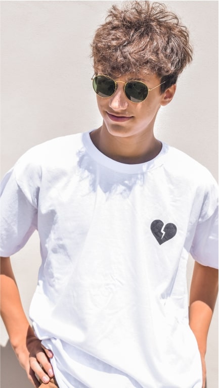 A young man posing in sunglasses and a white t-shirt with a broken heart design on the left chest.