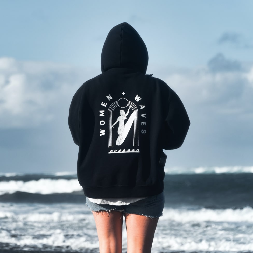 A woman on the beach wearing a black hoodie with a back design of a woman riding waves