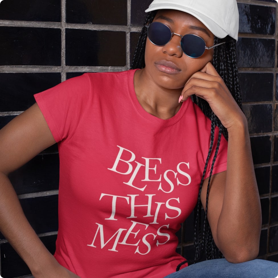 A woman wearing shades, a trucker's cap, and a pink t-shirt that says "Bless This Mess" in white letters