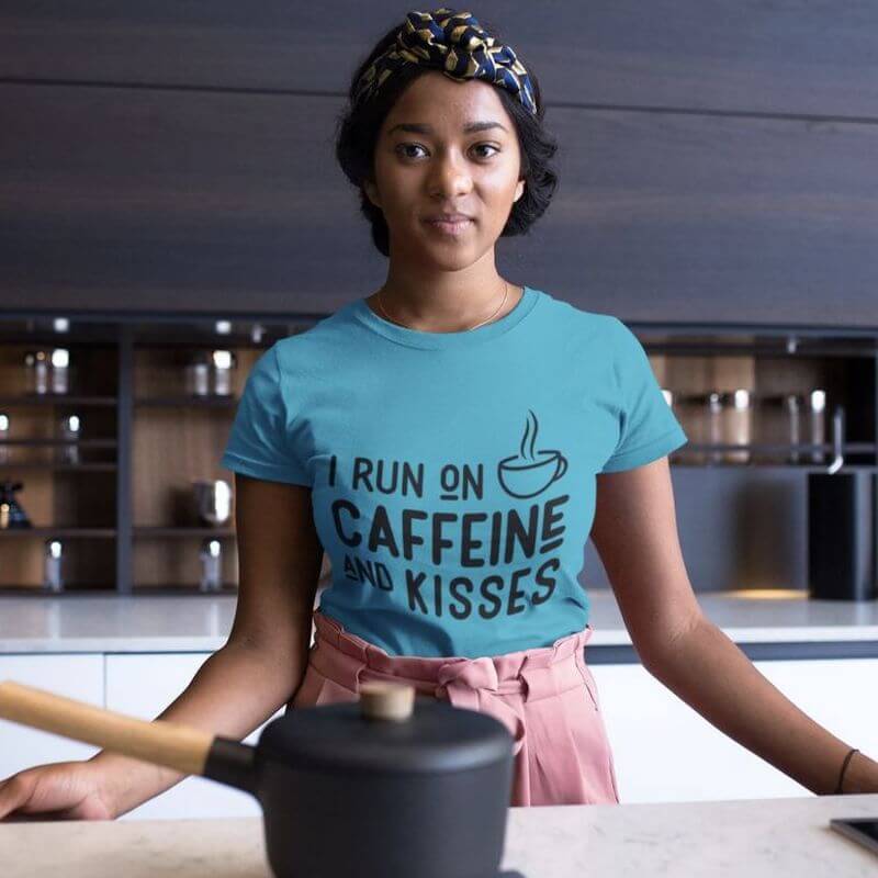 A woman posing in the kitchen in a blue t-shirt that says "I Run On Caffeine and Kisses"