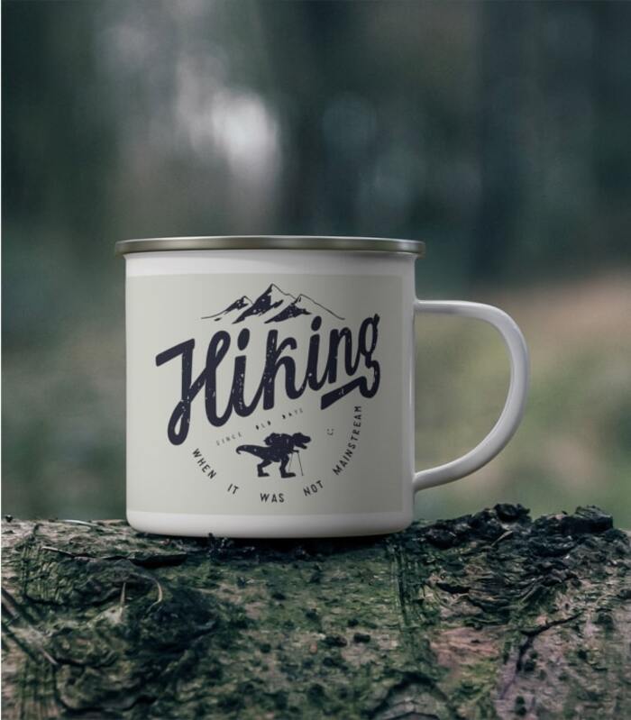 Enamel mug placed on a rock with the text “Hiking when it was not mainstream” around an image of a hiking dinosaur.
