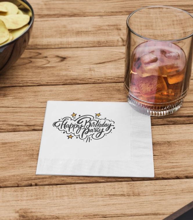 A white custom birthday napkin under a cup with a cocktail on a wooden table