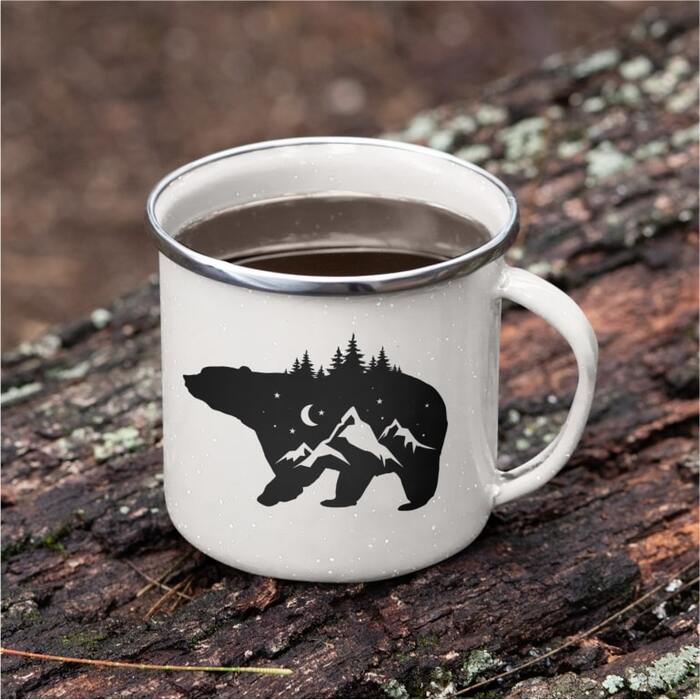 Enamel mug placed on a piece of wood with a stylized design of a bear filled with the night sky, mountains, and trees.