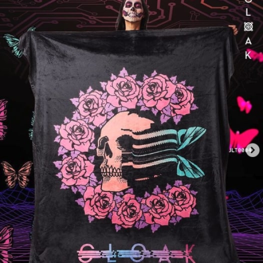 A woman holding up a large black blanket with a floral skull design and the brand name “Cloak” printed at the bottom.