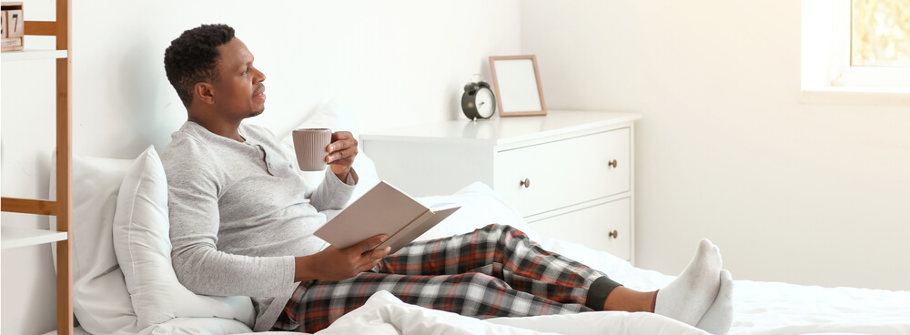 Man in bed with a book and a teacup, wearing custom pajamas with plaid pattern print on the pants and a plain gray shirt.