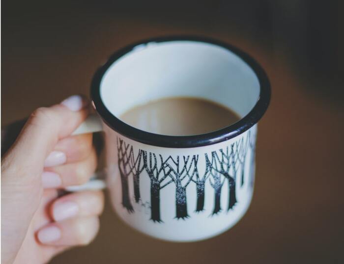 Person holding a white enamel mug with a design of black trees printed on it.