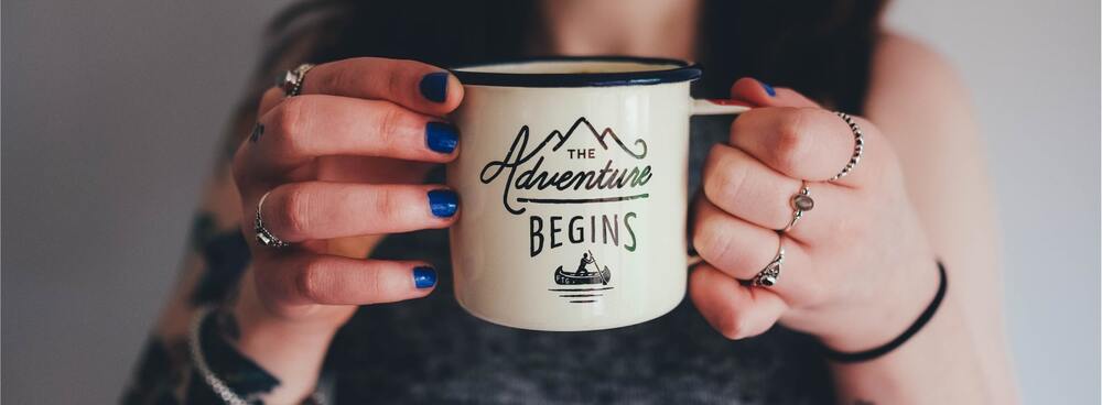 White camping mug with a stylized design of mountains and a person in a canoe, with the text “The Adventure Begins.”
