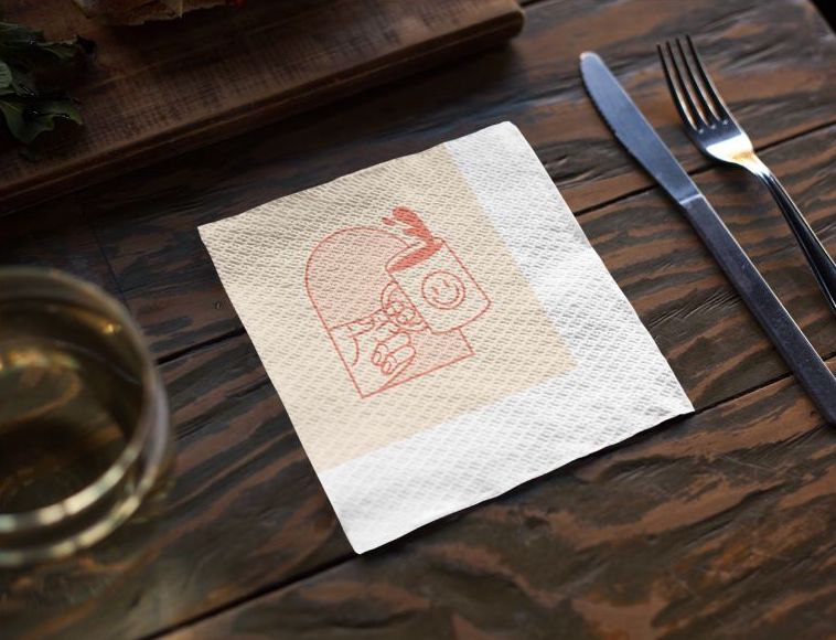 A small white paper napkin customized with a red smiling mug over a dark wooden table beside a fork and spoon