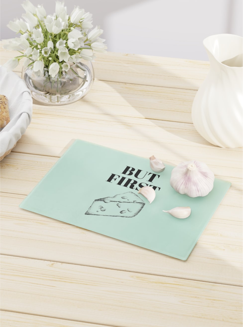 A glass cutting board customized with a sliced cheese design and the “But First” phrase over a wooden table