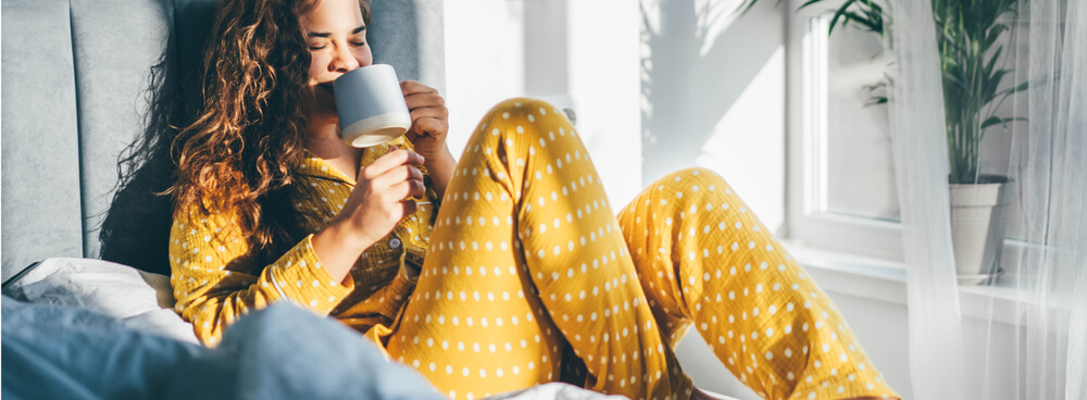 Woman drinking tea in bed, wearing a yellow pajama set with a white polka dot pattern.
