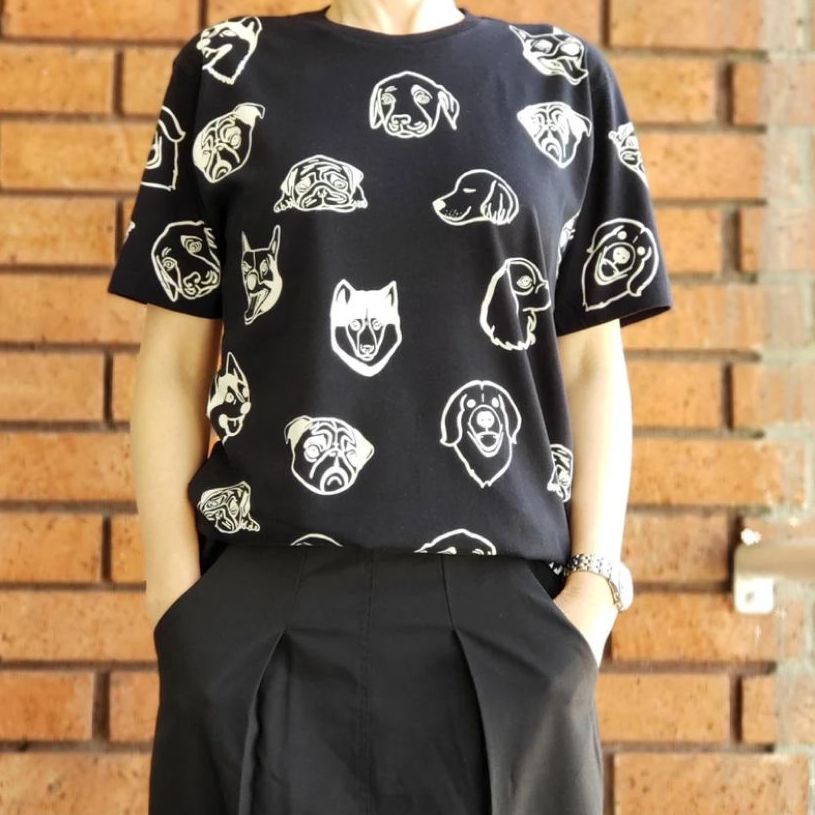 A woman standing in front of a red brick wall wearing a black all-over-printed t-shirt with a repeating white cartoon dog head pattern.