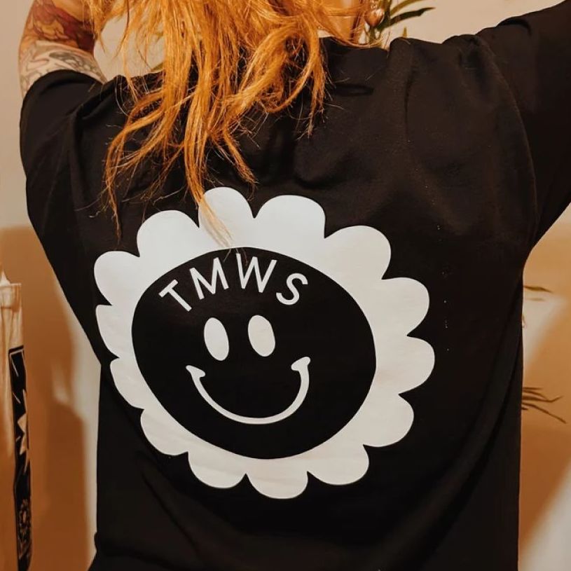 A person wearing a black t-shirt with an oversized white logo that looks like an abstract flower with the letters TMWS inside it and a smiley face below.