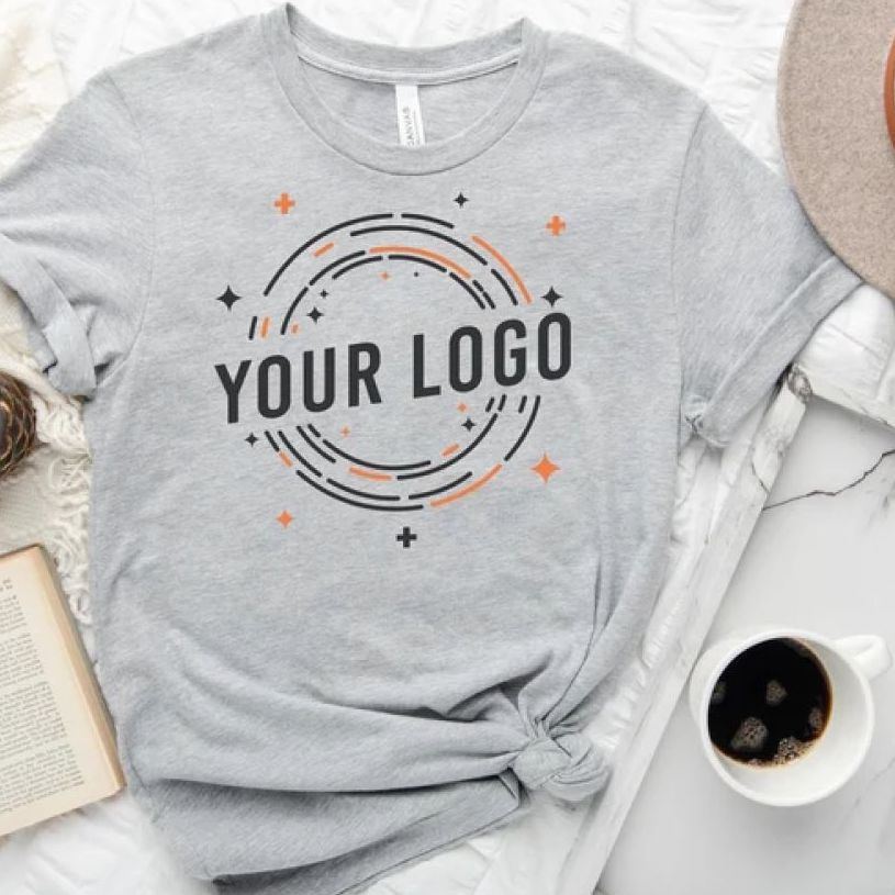 A light gray melange t-shirt that has an example logo illustration in black and orange laid flat on a surface with a cup of coffee beside it.