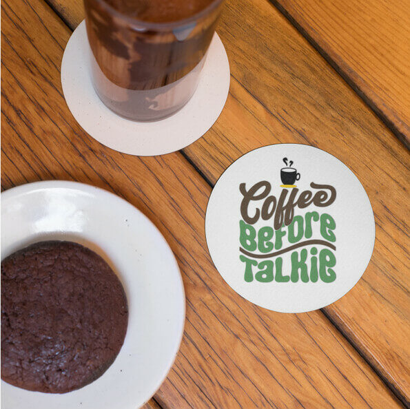 Round personalized coaster next to a muffin with the text “Coffee before talkie.”
