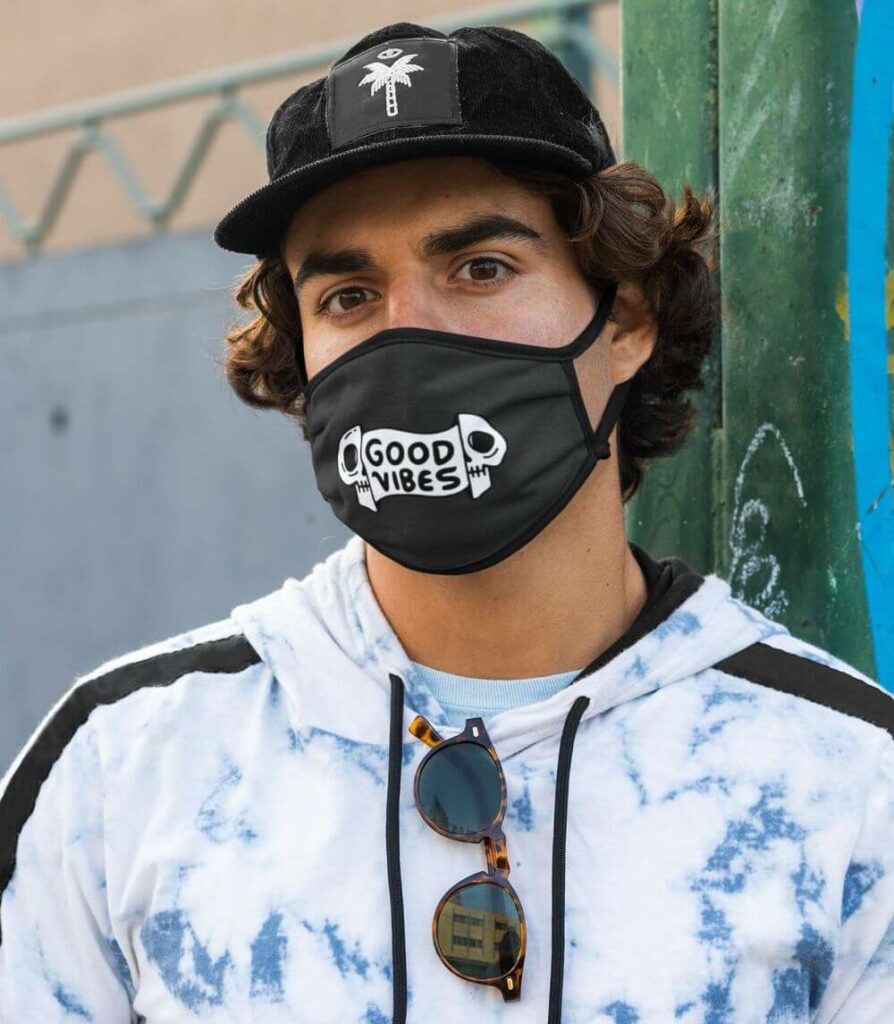 A man posing in a sublimed hoodie, cap with a palm tree design, sunglasses, and a custom mask that says "Good Vibes"