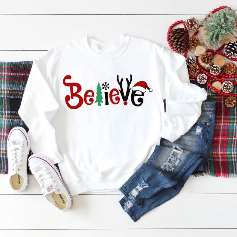 A white sweatshirt laid flat in a Christmassy setting with the words “Believe” printed in festive colors and motifs and a Santa's hat in the letter “E.”