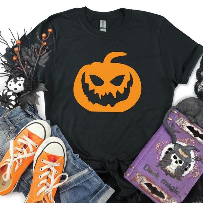 A black t-shirt laid flat in a Halloween setting with an orange cartoon jack-o-lantern printed on the chest.