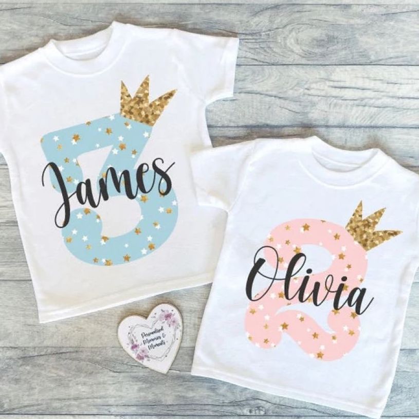 Two white toddler t-shirts with numbers “2” and “3,” as well as names James and Olivia and little gold crowns printed on them.