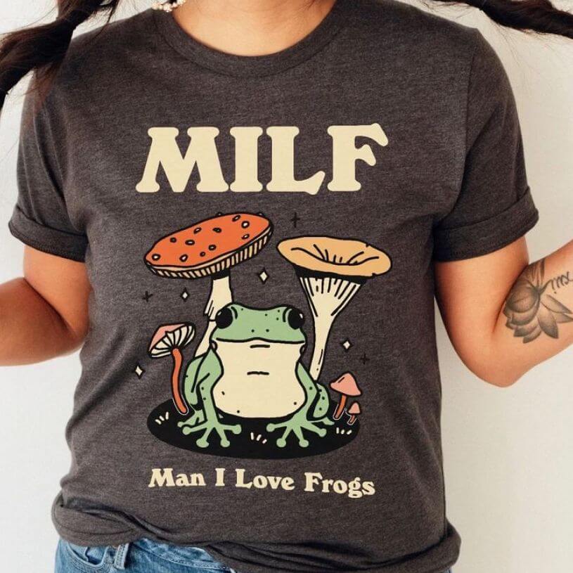 Dark gray t-shirt with a huge red, green, and beige colored print of mushrooms and a frog with the letters MILF on top and “Man I Love Frogs” on the bottom.