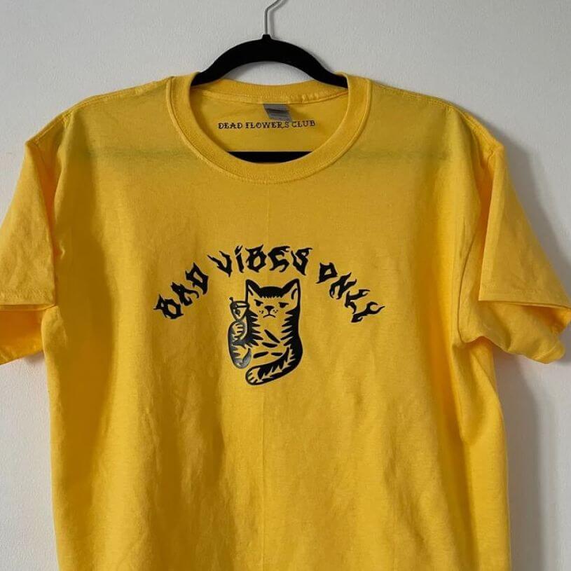 Yellow t-shirt with a “Bad Vibes Only” writing in black and a drawing of a grumpy cat drinking a martini.