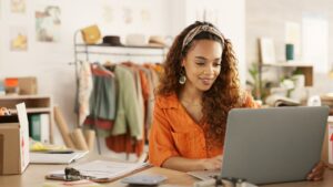 A woman working on a laptop with merchandise in the background.