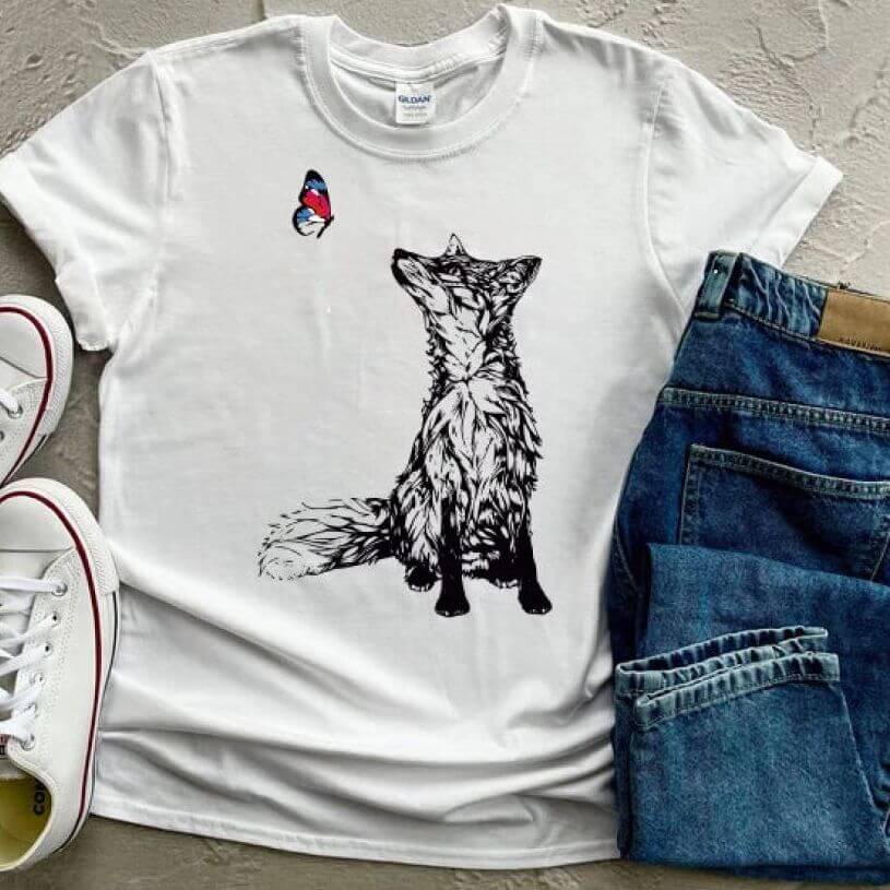 A white t-shirt laid flat beside a pair of jeans. The t-shirt has a drawing of a fox in black looking at a blue and red butterfly.