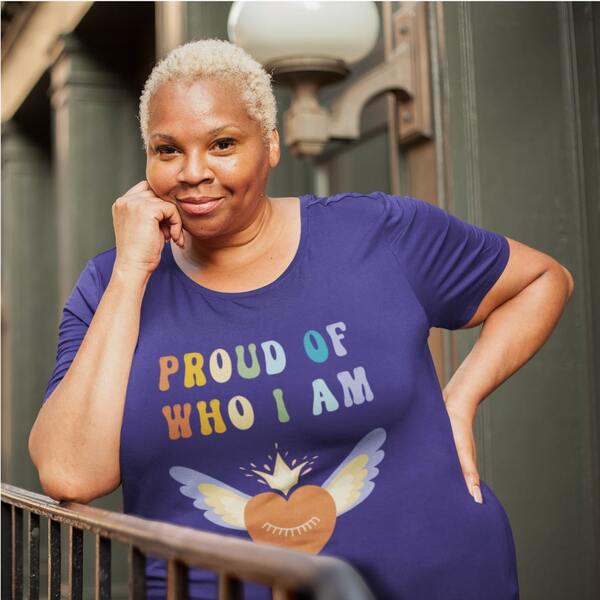 Lifestyle photo of a model leaning against a railing, posing in a custom t-shirt with the slogan “Proud of Who I Am” and the image of a heart with wings printed on the front.