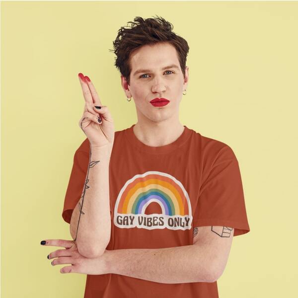 Model signals a peace movement hand sign while posing in a custom t-shirt designed with a rainbow above the slogan “Gay Vibes Only” printed on the chest.