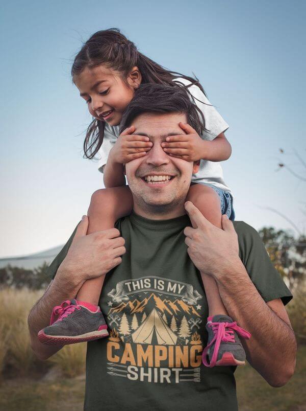 A dad holding his daughter on his shoulders, enjoying camping in a themed custom shirt that says, “This is my camping shirt.”